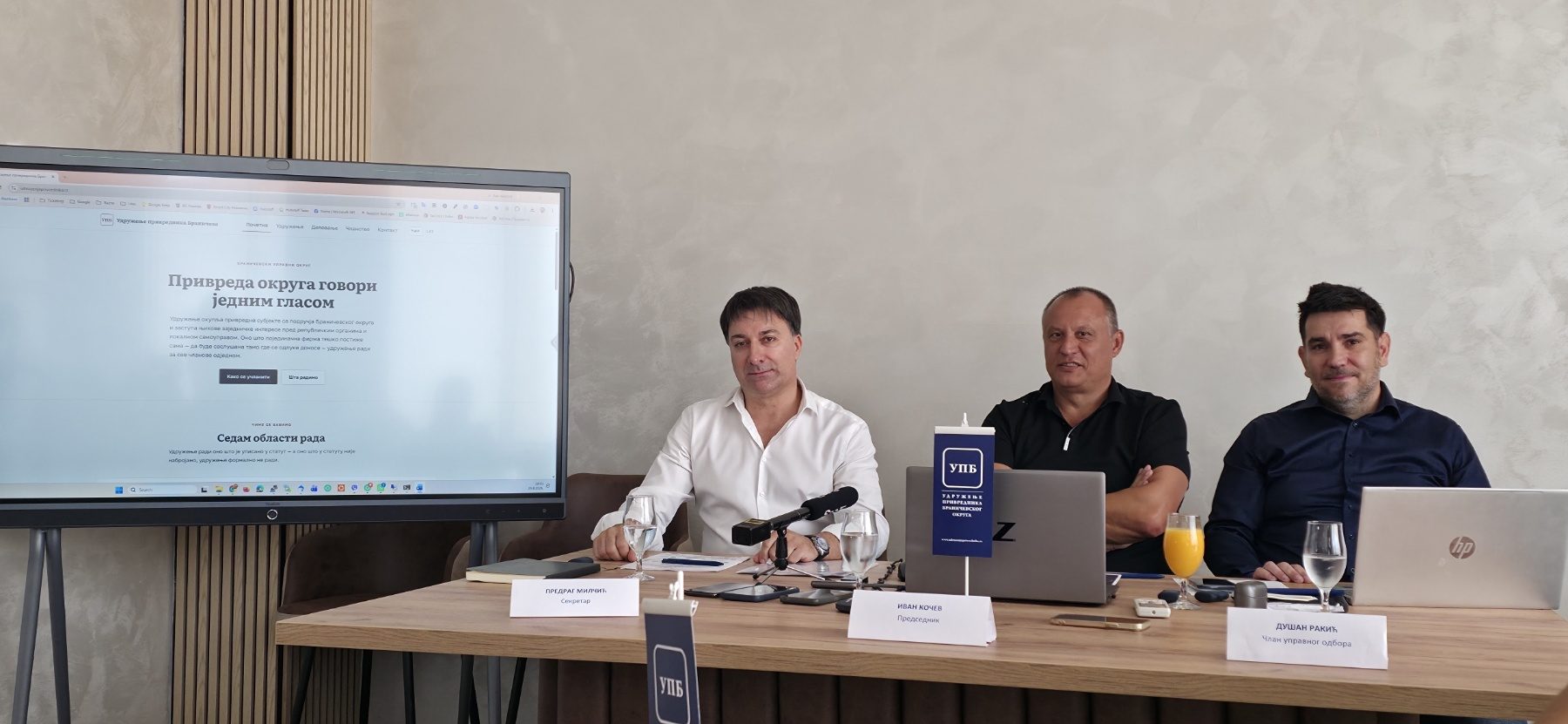 Predrag Milčić, Ivan Kočev i Dušan Rakić za stolom na konferenciji, uz zastavicu udruženja i sajt udruženja na ekranu